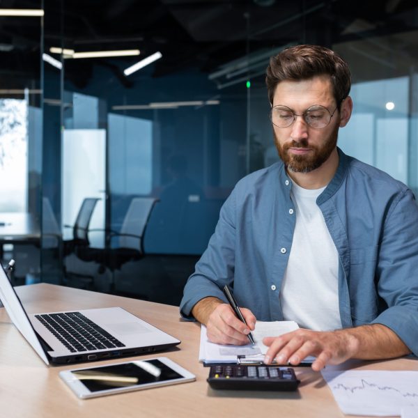 Serious and focused financier accountant on paper work inside office, mature man using calculator and laptop for calculating reports and summarizing accounts, businessman at work in casual clothes.
